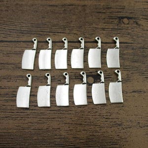 May include: A set of 15 silver-toned metal charms shaped like butcher knives. The charms are all the same size and shape, and they have a slightly distressed finish.