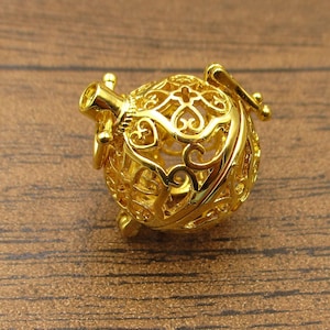 Op de afbeelding: Een gouden metalen bedel met een delicaat, ingewikkeld ontwerp. De bedel is bolvormig met een hol centrum en een kleine opening aan de zijkant. Het is een populaire keuze voor het maken van sieraden en kan worden gebruikt om kettingen, armbanden en oorbellen te maken.
