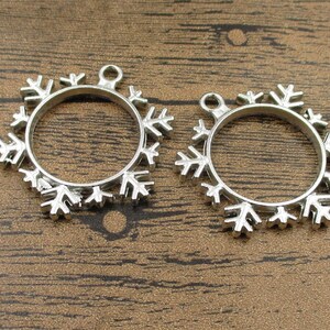 May include: Two silver snowflake-shaped pendant frames with a circular opening in the center. The frames are on a wooden surface.