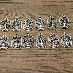 Puede incluir: Un conjunto de 12 dijes de metal plateado con un diseño de árbol de la vida. Cada dije es de forma rectangular con un diseño detallado e intrincado de un árbol con ramas y hojas.