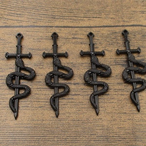 Puede incluir: Cinco colgantes de espada de metal negro con un diseño de serpiente. Las espadas tienen una guarda cruzada y una punta puntiaguda. Las serpientes están envueltas alrededor de las espadas y tienen un diseño detallado.