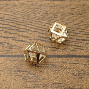 Puede incluir: Dos colgantes geom&eacute;tricos en forma de jaula de color dorado con inserciones de vidrio transparente. Los colgantes est&aacute;n sobre una superficie de madera marr&oacute;n.
