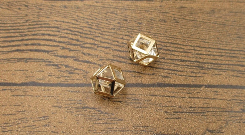 Puede incluir: Dos colgantes geom&eacute;tricos en forma de jaula de color dorado con inserciones de vidrio transparente. Los colgantes est&aacute;n sobre una superficie de madera marr&oacute;n.