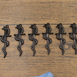 Puede incluir: Siete colgantes de metal negro con forma de serpiente enroscada alrededor de una daga. Los colgantes son todos del mismo tamaño y diseño.