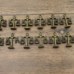 Puede incluir: Un conjunto de dijes de balanza de color bronce, cada uno con un diseño de barra de equilibrio y pesas colgantes. Los dijes están dispuestos sobre una superficie de madera, ideales para la creación de joyas o proyectos de artesanía.