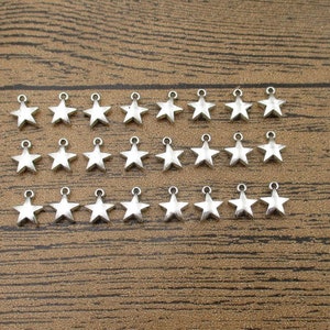 May include: A collection of 30 silver-toned star-shaped charms with a loop at the top. The charms are arranged in three rows of ten on a wooden surface.