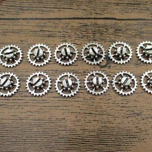 Puede incluir: Doce engranajes de metal plateado con un tornillo en el centro. Los engranajes están dispuestos en círculo sobre una superficie de madera marrón.