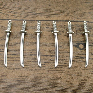 Könnte beinhalten: Sechs silberfarbene Katana-Anhänger mit detaillierten Griffen und gebogenen Klingen. Jeder Anhänger hat oben eine kleine Schlaufe zum Befestigen an Schmuck oder anderen Bastelarbeiten. Die Anhänger sind auf einer Holzoberfläche angeordnet.