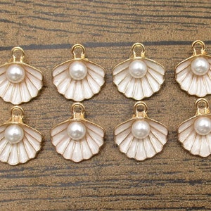 Op de afbeelding: Acht gouden schelpbedels met wit emaille en een parel in het midden. De bedels zijn perfect voor het maken van sieraden of om een ​​zee-achtige charme toe te voegen aan uw knutselwerk.