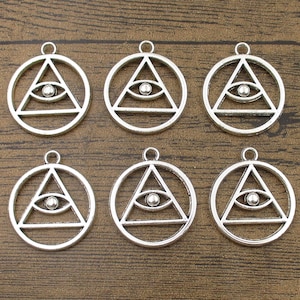 Puede incluir: Seis abalorios de metal plateado con un ojo que todo lo ve dentro de un triángulo, rodeado de un círculo. Los abalorios son perfectos para la elaboración de joyas o manualidades.