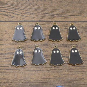 May include: Eight ghost-shaped charms arranged in two rows. Each charm features a smooth black enamel finish with two small white circular eyes and a gold-toned metal border. A small circular loop is attached to the top of each ghost figure.