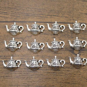 May include: Silver-colored teapot charms with the words "tea time" inscribed on the front. The charms are arranged on a brown wooden surface. These charms are ideal for jewelry making or crafting projects.