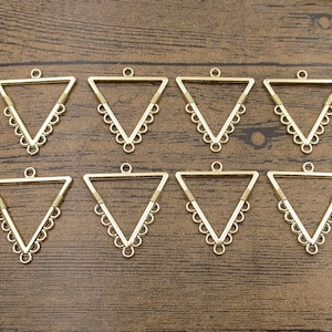 Op de afbeelding: Tien gouden driehoekige metalen hangerblanks met een decoratieve rand. Elke hanger heeft een lus aan de bovenkant om een ​​ketting of koord aan te bevestigen.