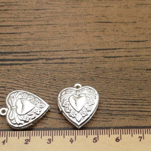 May include: Two silver heart-shaped locket charms with ornate floral designs. The charms are approximately 3 inches long and are shown on a wooden surface with a ruler for scale.