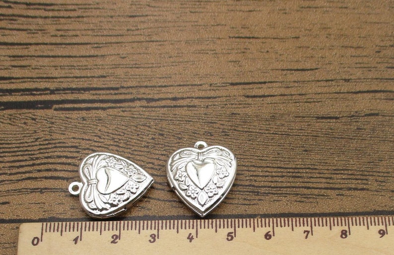May include: Two silver heart-shaped locket charms with ornate floral designs. The charms are approximately 3 inches long and are shown on a wooden surface with a ruler for scale.