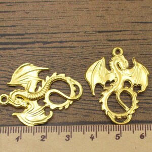 May include: Two gold-colored metal dragon charms. The dragon on the left is curled up with its tail in its mouth. The dragon on the right has its wings spread out. Both charms have a hole at the top for attaching to a necklace or bracelet.