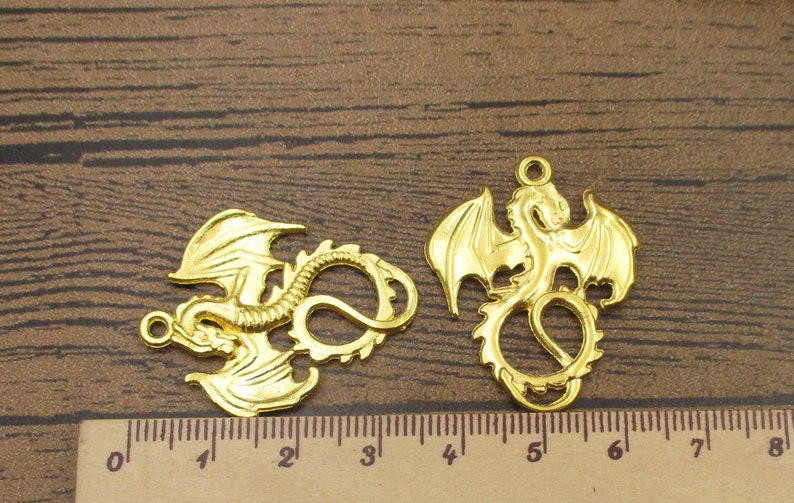 May include: Two gold-colored metal dragon charms. The dragon on the left is curled up with its tail in its mouth. The dragon on the right has its wings spread out. Both charms have a hole at the top for attaching to a necklace or bracelet.