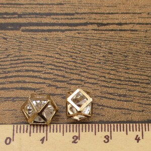 Puede incluir: Dos formas geom&eacute;tricas de metal dorado con acentos de cristal transparente. Las formas miden aproximadamente 2,5 cm y 3,8 cm de di&aacute;metro. Las formas est&aacute;n sobre una superficie de madera marr&oacute;n con una regla para la escala.