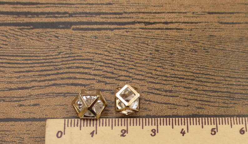 Puede incluir: Dos formas geom&eacute;tricas de metal dorado con acentos de cristal transparente. Las formas miden aproximadamente 2,5 cm y 3,8 cm de di&aacute;metro. Las formas est&aacute;n sobre una superficie de madera marr&oacute;n con una regla para la escala.
