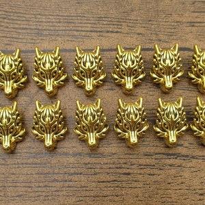 May include: Twelve gold-coloured wolf head charms are displayed. Each charm has a detailed wolf face design with defined features and a textured surface. The charms are arranged on a wooden surface.