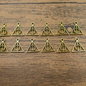May include: A set of 12 gold-toned metal charms shaped like triangles with a circle inside. The charms are all the same size and shape, and they are arranged in a row on a wooden surface.