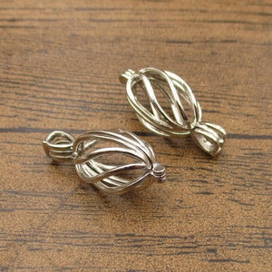 Könnte beinhalten: Zwei silberne Metallkäfig-Anhänger zum Halten von Perlen oder anderen kleinen Charms. Die Käfige sind oben offen und haben eine Schlaufe zum Anbringen an einer Kette oder Schnur.