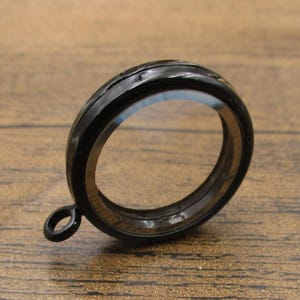 May include: A black curtain ring with a small loop attached. The ring is made of a glossy material and has a circular shape. The background is a wooden surface.