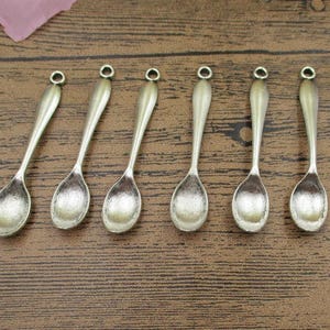 May include: Seven antique silver-toned spoon charms with a rounded bowl and a loop at the top.