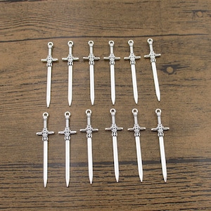 Puede incluir: Un conjunto de 18 dijes de espada plateados. Los dijes están detallados con diseños intrincados y tienen un bucle en la parte superior para sujetarlos a joyas u otras manualidades.