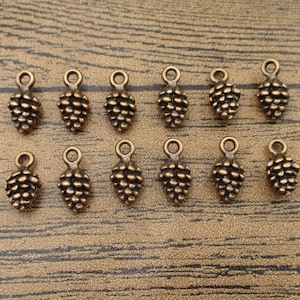 Puede incluir: Un conjunto de 12 abalorios de metal de color dorado antiguo en forma de piña con un bucle para colgar. Los abalorios están dispuestos en dos filas sobre una superficie de madera.