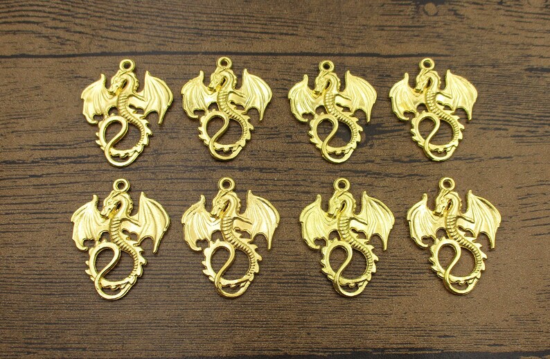 May include: Eight gold-colored metal dragon charms with wings outstretched. The charms are detailed and have a three-dimensional appearance.