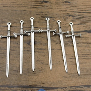 May include: Seven silver-toned sword charms with a cross guard and a pointed tip. The charms are arranged in a row on a wooden surface.