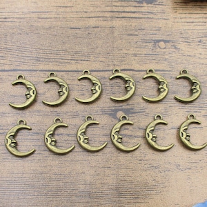 May include: A set of bronze-toned crescent moon charms, each featuring a face design. These charms have a small loop for attaching to jewellery or crafts. They are displayed on a wooden surface.