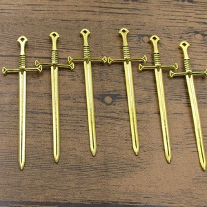 May include: Seven gold-colored miniature swords with cross guards. The swords are arranged in a row on a wooden surface.
