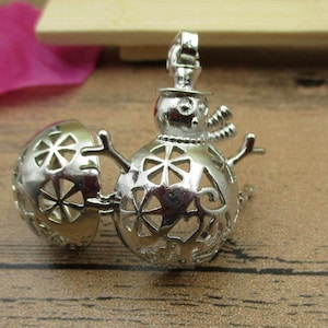 May include: Silver snowman charm with a hollow, openwork design. The charm is made of two halves that open to reveal a small space inside. The snowman is wearing a hat and has a small, round face.