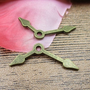 May include: Two antique gold-toned metal clock hands with pointed tips. The hands are arranged in a cross shape.