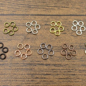 Può includere: Assortimento di anelli aperti di vari colori, tra cui oro, argento, nero e rame. Ogni colore è disposto in un cerchio con sette anelli aperti.
