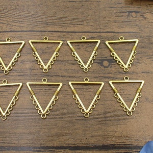 May include: Eight gold triangle-shaped metal pendant charms with a decorative scalloped edge. Each charm has a loop at the top for attaching to a necklace or bracelet.