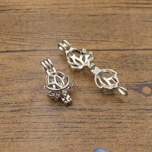 Puede incluir: Dos colgantes de jaula de metal plateados con diseños intrincados. Las jaulas están abiertas y parecen estar diseñadas para contener una pequeña perla u otro objeto.