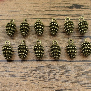 May include: Twelve antique gold-toned metal pine cone charms with a loop for hanging. The charms are arranged in two rows on a wooden surface.
