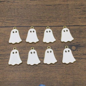 May include: Eight small ghost-shaped charms featuring white enamel surfaces with two black dots for eyes. Each charm has a gold-toned metal border and a small circular loop at the top for attachment, arranged in two rows of four identical pieces.