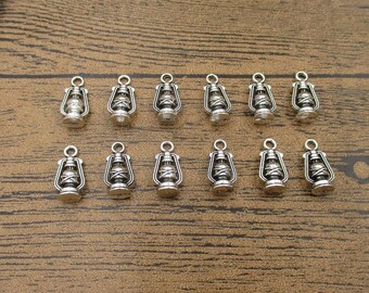May include: A set of 12 silver-toned metal charms shaped like oil lamps. The charms have a detailed design and are perfect for adding a vintage touch to jewelry or crafts.