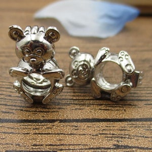 Puede incluir: Dos abalorios de plata con forma de osito de peluche. Los abalorios están detallados con una cara redonda y orejas pequeñas.