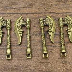 Könnte beinhalten: Satz mit vier antiken goldfarbenen Axt-Anhängern mit filigranen Details. Jeder Anhänger zeigt eine geflügelte Axtkopf mit einem dekorativen Griff.