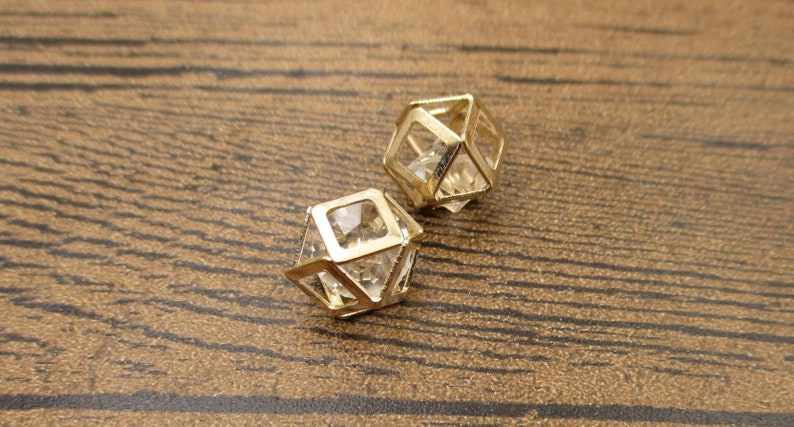 Puede incluir: Dos pendientes geom&eacute;tricos de tono dorado con detalles de cristal transparente. Los pendientes tienen forma de cubos con los lados abiertos.