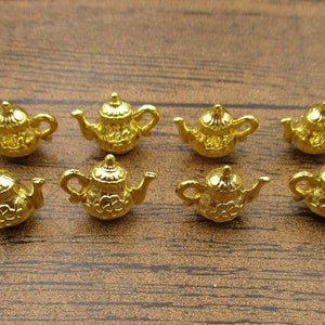 Könnte beinhalten: Acht goldene Miniatur-Teekannen mit verzierten Details. Die Teekannen sind in zwei Reihen zu je vier auf einer Holzoberfläche angeordnet.