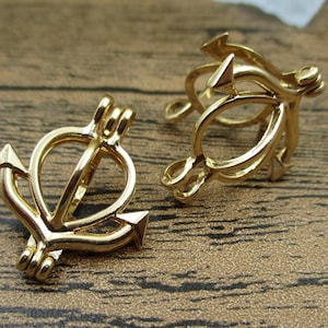 Puede incluir: Dos colgantes de metal dorado en forma de ancla con una pequeña abertura en la parte superior. Los colgantes están sobre una superficie de madera.