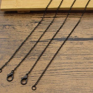 Può includere: Quattro collane a catena nera con chiusura a moschettone. Le catene sono di diverse lunghezze e sono disposte su una superficie di legno.