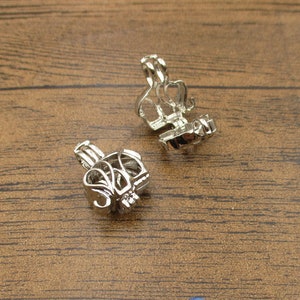 Puede incluir: Dos colgantes de jaula de metal plateados con un diseño decorativo. Las jaulas están abiertas y parecen estar diseñadas para contener un objeto pequeño, como una perla o un amuleto.