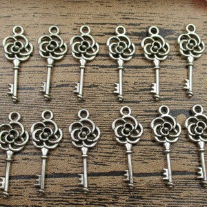 May include: A collection of antique silver-colored keys. Each key features a decorative floral design at the top and a traditional key shape. The keys are arranged on a wooden surface, ideal for jewelry making or craft projects.